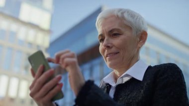 Kendinden emin yaşlı bir kadın modern şehir ortamında akıllı telefonuyla iletişim kurar, odaklanarak gezer ya da kayar ve kolaylıkla.