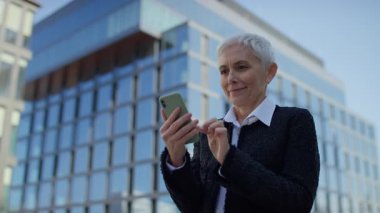 Modern bir şehir ortamında akıllı telefonundan olumlu haberler okurken iş kıyafetleri içindeki mutlu yaşlı kadın heyecanlanmış görünüyor..