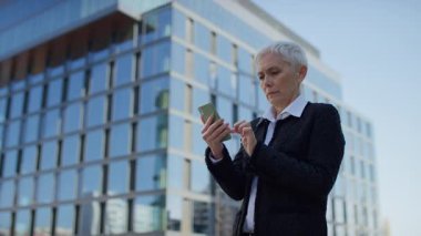 İş kıyafetleri içinde sıkıntılı yaşlı bir kadın akıllı telefonuna alarmla bakar. Beklenmedik ya da rahatsız edici haberlere tepki verir..