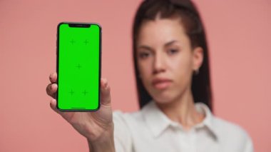 Genç bir kadın, yeşil ekran maketi ile dikey bir akıllı telefon tutarak kolunu ileri uzatıyor, yumuşak pembe bir arka plana doğru..