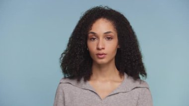 Kıvırcık saçlı genç bir kadının portresi tasvip etmeksizin kafasını sallıyor, düz mavi bir stüdyo arka planına karşı..