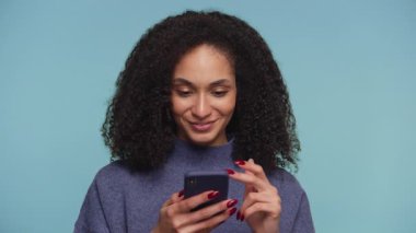 Akıllı telefonuna bakarken gülümseyen ve tezahürat yapan kıvırcık saçlı neşeli genç kadın heyecan verici haberlere ya da başarıya tepki veriyor..