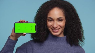 Mavi arkaplan önünde yeşil ekranlı akıllı bir telefonu tutarken, kıvırcık saçlı mutlu genç kadın onaylıyor..