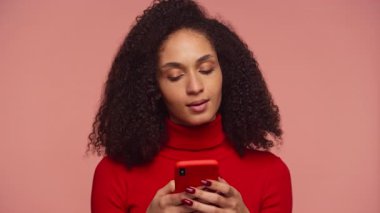 Sinirli, kıvırcık saçlı, telefonunda yoğun mesaj atan genç bir kadın, gözleri hayal kırıklığı içinde, pembe bir arka plana karşı kapalı..