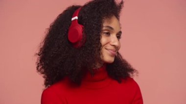 Kıvırcık saçlı, kırmızı kulaklık takan ve kırmızı kazaklı genç bir kadın. Pembe arka planda müzik dinlerken huzur içinde gülümsüyor..