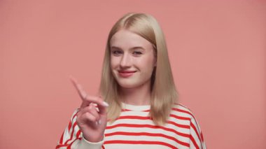 Gülümseyen sarışın kadın, neşeli bir uyarı jesti ile işaret parmağını kaldırıyor, pembe bir arka plana karşı neşeli bir tedbir taşıyor..