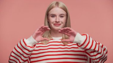 Kırmızı çizgili tişörtlü sarışın kadın sıcak bir şekilde gülümsüyor ve yumuşak pembe arka planda elleriyle kalp şekli oluşturuyor..