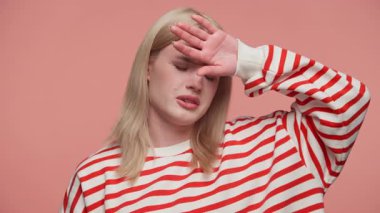 Çizgili tişörtlü sarışın kadın elini alnına dayayıp gözlerini kapatıyor. Görünürde iyi değil ya da hasta. Pembe arka planda..