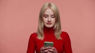 Kırmızı balıkçı yakalı sarışın kadın akıllı telefonuyla yazı yazarken aşağıya bakıyor, odaklanmış ve pembe bir arka plana yaslanmış..