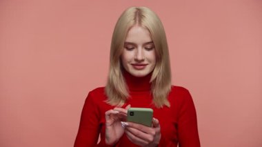 Kırmızı balıkçı yaka kazaklı sarışın kadın akıllı telefonuyla kayarken gülümsüyor, onaylayarak başını sallıyor ve çevrimiçi içerikten keyif alıyor..