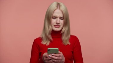 Kırmızı balıkçı yakalı sarışın kadın mesaj atarken kaşlarını çatıyor, telefonundaki hayal kırıklığı yaratan ya da üzücü bir mesaja duygusal tepki veriyor, pembe arka planda izole edilmiş..