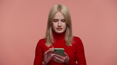 Kırmızı balıkçı yaka kazaklı sarışın kadın elinde akıllı bir telefon tutarken sinirli görünüyor. Pembe arka plandan alışveriş yaparken memnuniyetsizliğini dile getiriyor..