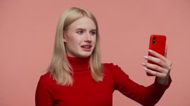 Kırmızı balıkçı yaka kazaklı sarışın kadın video görüşmesi sırasında gülümsüyor ve konuşuyor. Önünde pembe arka plana karşı akıllı telefonu tutuyor..