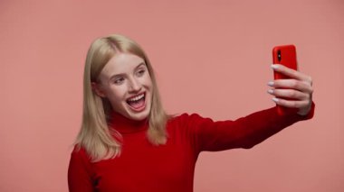 Kırmızı balıkçı yaka kazaklı neşeli sarışın kadın akıllı telefon tutuyor ve pembe arka planda eğlenceli bir selfie çekerken geniş bir gülümseme sergiliyor..