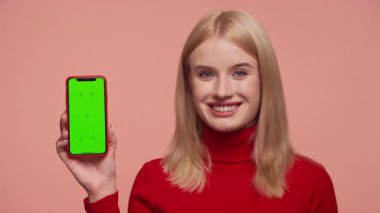 Kırmızı balıkçı yaka kazaklı, elinde yeşil ekranlı akıllı telefon tutan gülümseyen sarışın kadın, pembe arka planda izole edilmiş mobil bir uygulama ile olumlu eleştiri veya memnuniyeti sembolize ediyor..