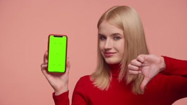 Kırmızı balıkçı yaka kazaklı sarışın kadın yeşil ekranlı akıllı telefonu gösteriyor ve bir uygulamadan hayal kırıklığı yaratıyor, pembe arka planda izole edilmiş.