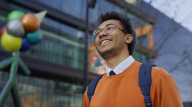 Sırt çantalı gülümseyen genç adam modern akademik binanın önünde duruyor, atmosferin tadını çıkarıyor ve iyimserlikle yukarı bakıyor..