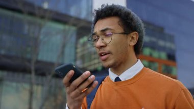 Sırt çantalı genç adam akıllı telefona konuşuyor, ses asistanıyla iletişim kuruyor ya da modern kampüs binalarının yakınında sesli mesaj kaydediyor..