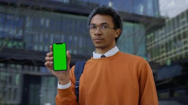 Turuncu tişörtlü hoşnutsuz genç adam yeşil ekranlı akıllı telefonu tutarken başını sallıyor hayır, dışarıda tatminsizliğini dile getiriyor..
