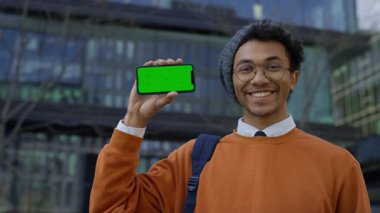 Turuncu tişörtlü mutlu genç adam yeşil ekranlı akıllı telefonu tutuyor ve modern binaların yakınındaki açık havada memnuniyetini belirterek onaylıyor..