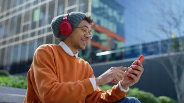 Kulaklık takan gülümseyen genç adam modern bir şehir ortamında dışarıdaki merdivenlerde otururken bir video görüşmesi sırasında selam veriyor..
