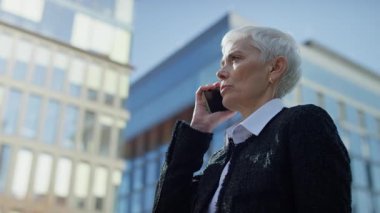 Resmi kıyafetli kıdemli profesyonel bir kadın modern bir iş bölgesinde açık havada ciddi bir telefon görüşmesi yapıyor..