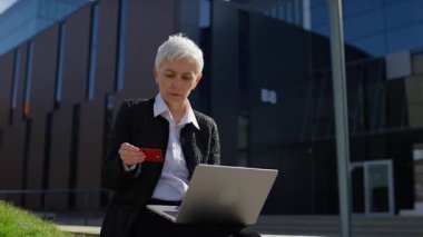 Kredi kartı tutan ve dizüstü bilgisayara endişeli bakan yaşlı bir kadın, çevrimiçi bir işlemin başarısız olduğunu veya geri çevrildiğini gösteriyor.