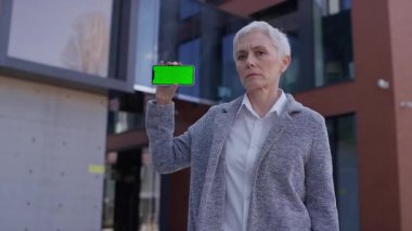 Mature woman with serious expression holding smartphone with green screen, symbolizing customer dissatisfaction and negative feedback on app or service.