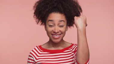 Kıvırcık saçlı, başarıyı kutlayan, kırmızı çizgili gömleğiyle pembe arka planda gülümseyen mutlu bir genç kadın..