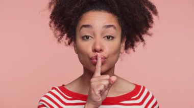 Susturucu bir jest yaparak parmaklarını dudaklarına dayayan genç bir kadın. Sessizlik ya da gizlilik işareti yapıyor. Pembe arka planda kırmızı çizgili gömlek giyiyor..