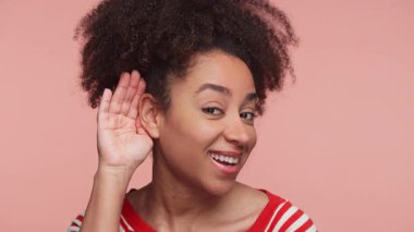 Çizgili tişörtlü genç bir kadın tuhaf bir gülümsemeyle el ele tutuşuyor, pembe arka planda izole edilmiş, dinlemeyi ya da dikkat etmeyi sembolize ediyor..