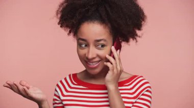 Kırmızı çizgili tişörtlü genç bir kadın cep telefonuyla konuşuyor. Konuşma sırasında gülümsüyor ve el kol hareketi yapıyor. Pembe arka planda izole edilmiş..