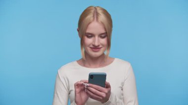 Neşeli genç bir kadın elinde akıllı telefon tutarken heyecanlı görünüyor. Mavi arka plana karşı büyük bir heyecan ve sürprizle tepki veriyor..