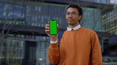 Turuncu kazaklı genç adam yeşil ekranlı bir akıllı telefon gösteriyor ve bir uygulamadan memnun olmadığını öne sürüyor..