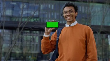 Gözlüklü mutlu genç adam yeşil ekranlı akıllı telefonu gösteriyor. Dijital bir ürünün onaylanması ya da tanıtımı öneriliyor..