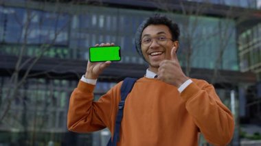 Turuncu kazaklı neşeli genç adam yeşil ekranlı akıllı telefon gösteriyor ve memnuniyeti veya onayını işaret ediyor..