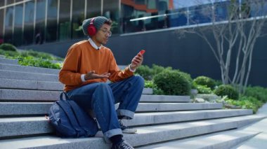 Kulaklık takan genç bir adam video konuşmasında modern açık hava basamaklarında oturan akıllı telefonunu kullanarak konuşuyor..