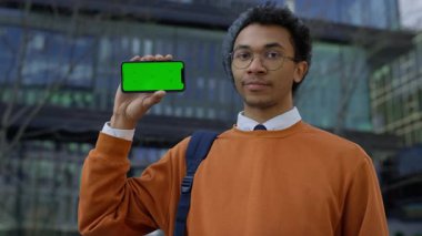 Turuncu kazaklı genç adam yeşil ekranlı akıllı telefonu tutarken başını sallıyor. Negatif eleştiri veya geri bildirim sinyali veriyor..