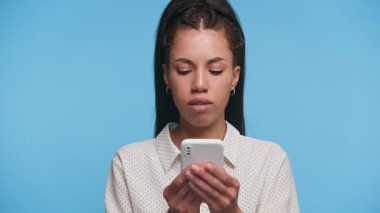 Beyaz gömlekli, gülümseyen genç bir kadın, akıllı telefondan olumlu haberler okuduktan sonra kameraya bakar, mavi arka planda durur..