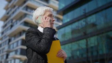 Kısa gri saçlı, kendinden emin yaşlı kadın elinde klasörle modern ofis binalarının önünde dikilirken akıllı telefonuyla konuşuyor..