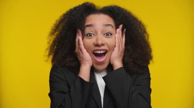 Heyecanlı, kıvırcık saçlı, şaşkınlık içinde yüzünü tutan genç bir kadın, parlak sarı bir arka plana karşı harika haberlere tepki veriyor..