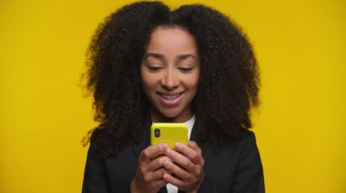 Sarı bir telefonla mesajlaşırken, sarı arka planda heyecan ve mutluluk gösteren kıvırcık saçlı genç bir kadın..