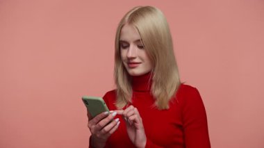 Kırmızı balıkçı yaka kazaklı sarışın kadın telefonuna bakıyor. Pembe yumuşak bir arka plana karşı neşe ve şaşkınlıkla tepki veriyor..