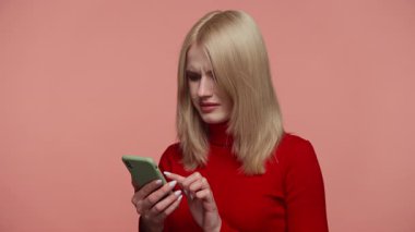 Kırmızı elbiseli sarışın kadın, pembe arka plandaki başarısız alıma tepki olarak, kafası karışmış ve hayal kırıklığına uğramış bir şekilde telefona bakıyor..