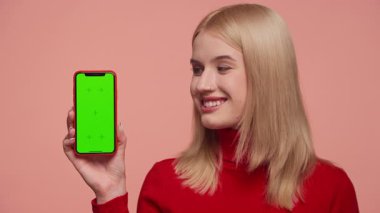 Kırmızı balıkçı yaka yakalı sarışın kadın elinde yeşil ekranlı akıllı telefon tutuyor. Uygulama veya pembe arka plandaki içerik modeli için ideal..