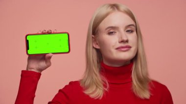 Kırmızı balıkçı yakalı sarışın kadın yeşil ekranlı telefonu elinde tutarken pembe arka plana karşı kafasını sallıyor..