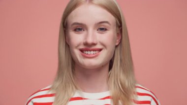 Sarışın kadın, kırmızı ve beyaz çizgili gömleğiyle sıcak bir şekilde gülümsüyor. Dost canlısı bir yakın çekim portresi için pembe bir arka plana poz veriyor..