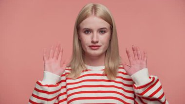 Çizgili, kırmızı ve beyaz kazaklı sarışın kadın her iki elini de kaldırıyor ya da pembe arka planda yumuşak bir jesti durduruyor..