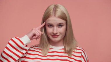 Kırmızı çizgili süveter giyen sarışın kadın şakağına hafifçe dokunurken gülümsüyor. Pembe arka plana karşı zekice ya da akıllıca bir hareket yapıyor..