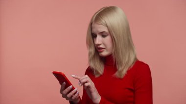 Kırmızı kazaklı sarışın kadın şok olmuş görünüyor. Akıllı telefondan rahatsız edici bir mesaj okuyor. Ağzı inançsızlıkla örtüyor, pembe arka planda izole edilmiş..